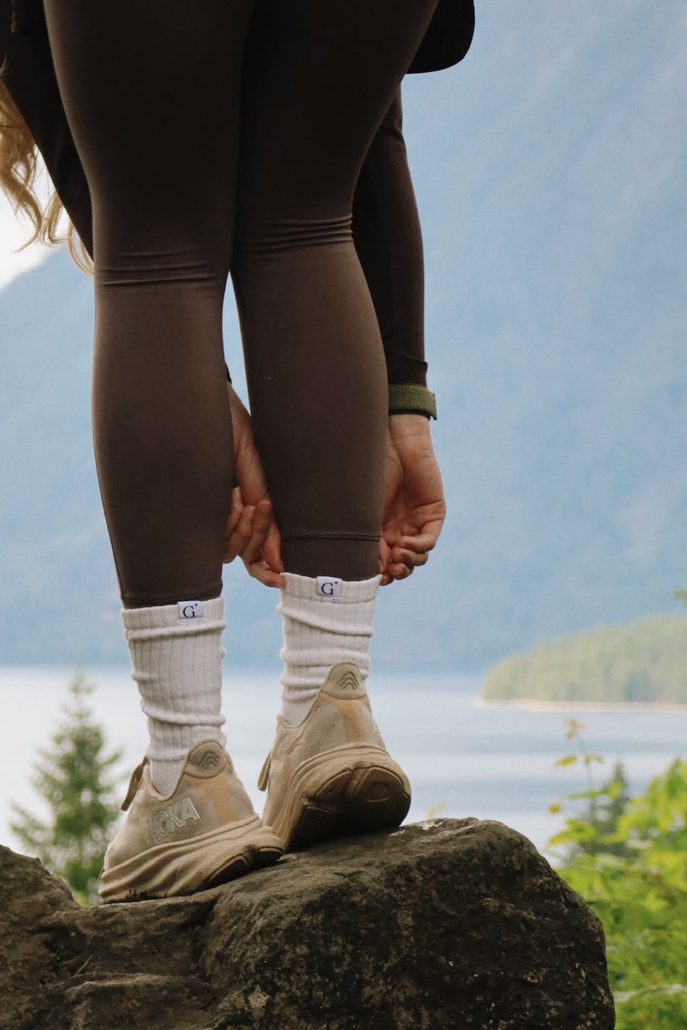 A person standing on a rock by a lake with mountains in the background, wearing brown leggings, white crew socks with a small blue logo, and beige hiking shoes.