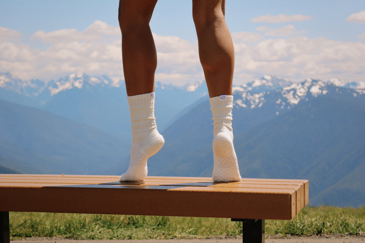 Person wearing white socks standing on a wooden bench with mountains in the background