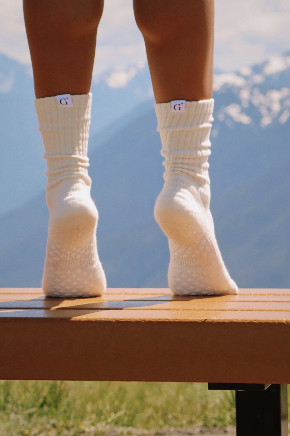 Person wearing white socks with a brand logo, standing on a wooden bench with mountains in the background.